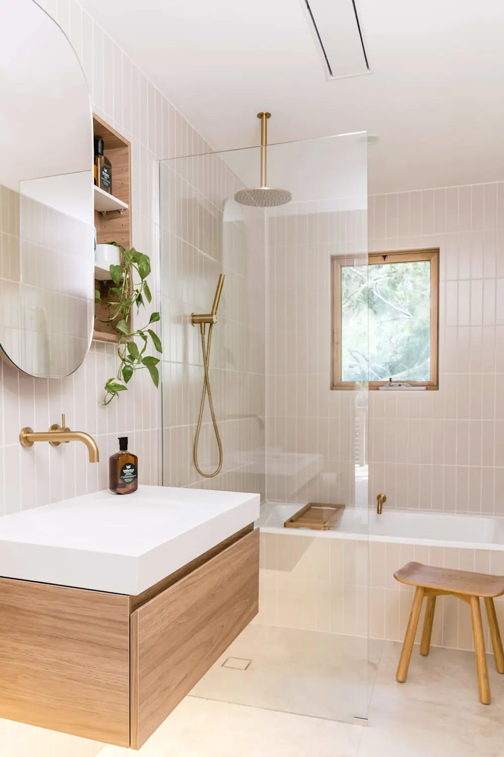 Modern bathroom with cream tiles, glass shower, wooden vanity, brass fixtures, and a small window overlooking greenery.