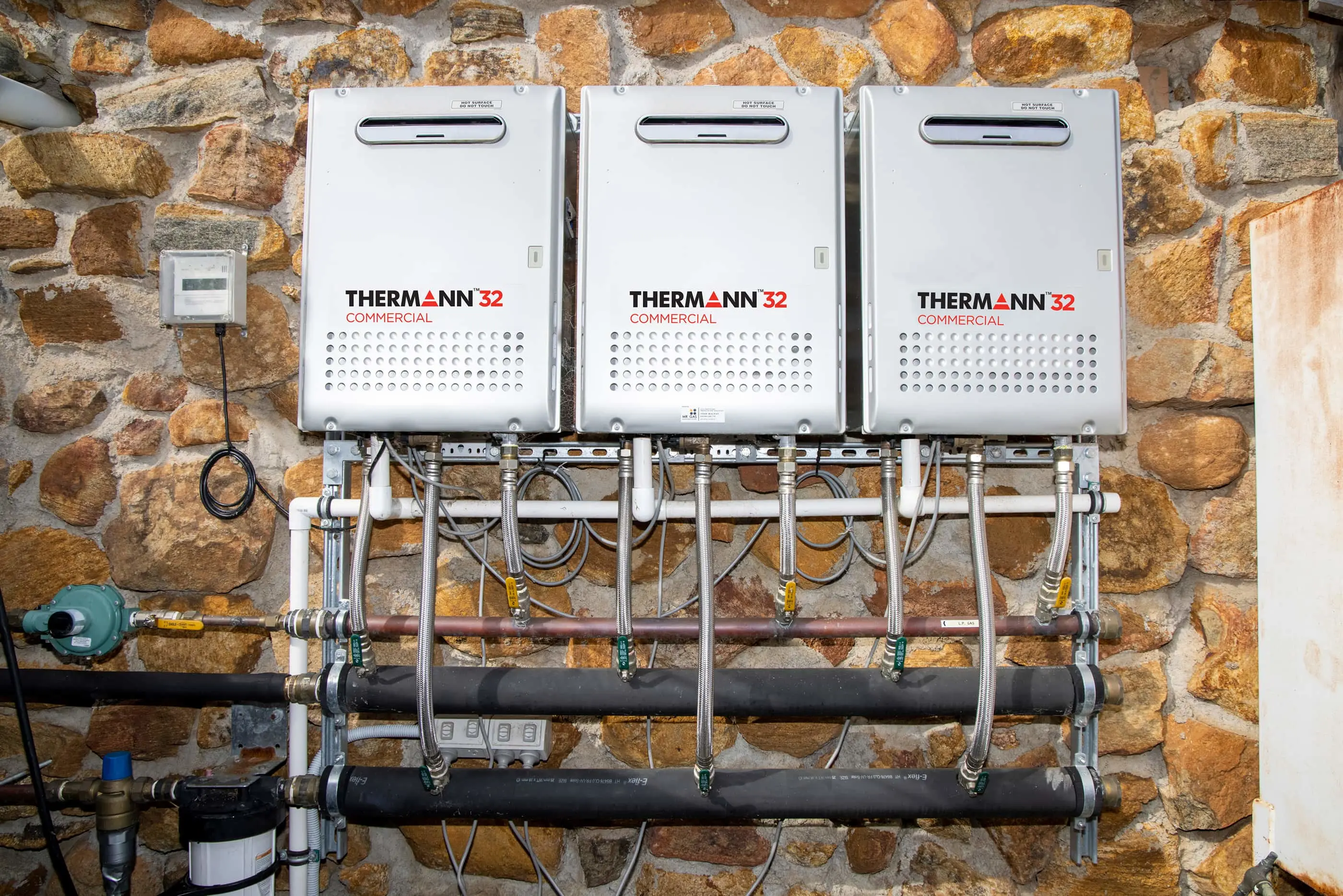 Three Thermann Commercial water heaters mounted on a stone wall with connected pipes and plumbing system.