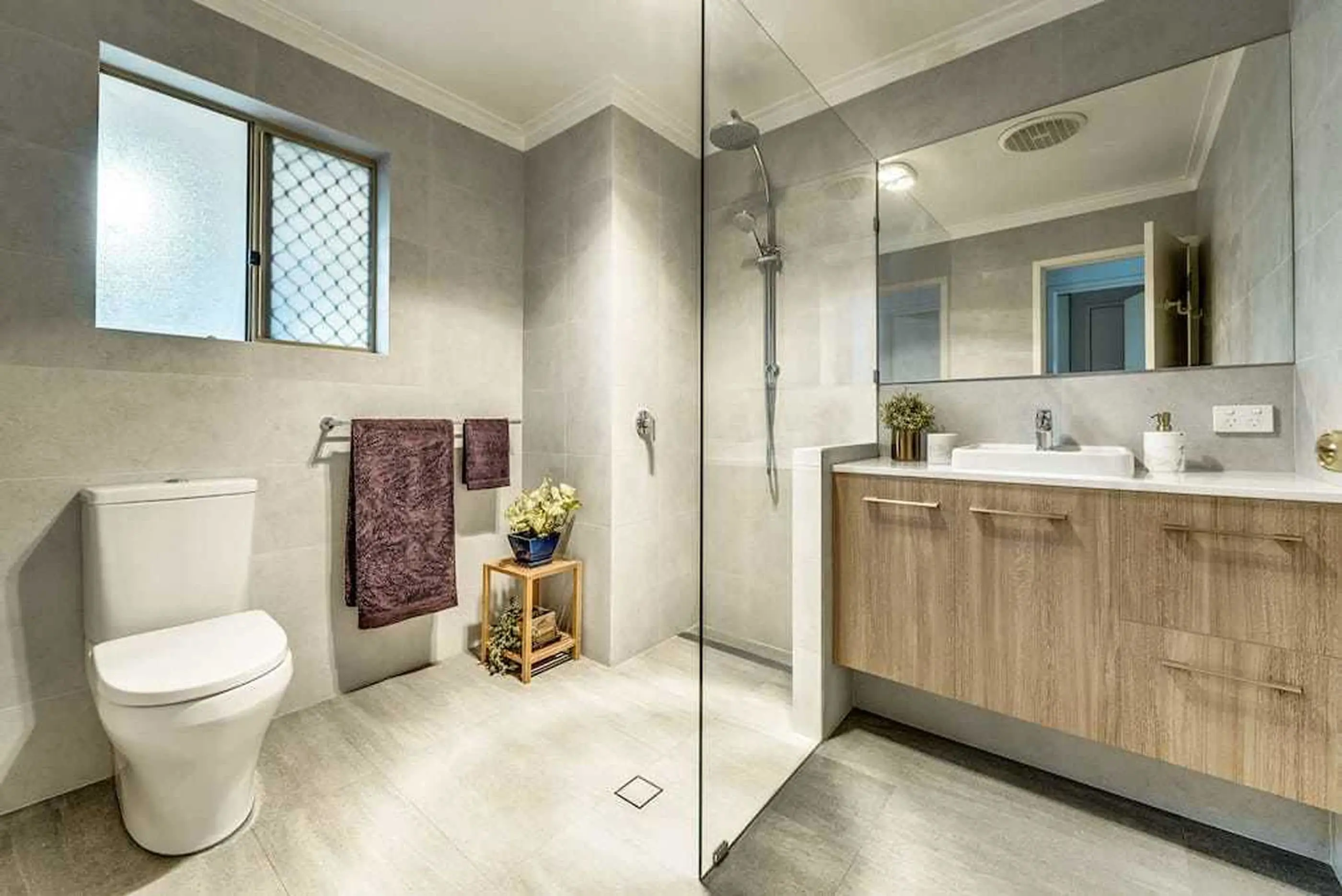 Modern bathroom with gray tiles, glass shower, wooden vanity, white toilet, and purple towels on rack.