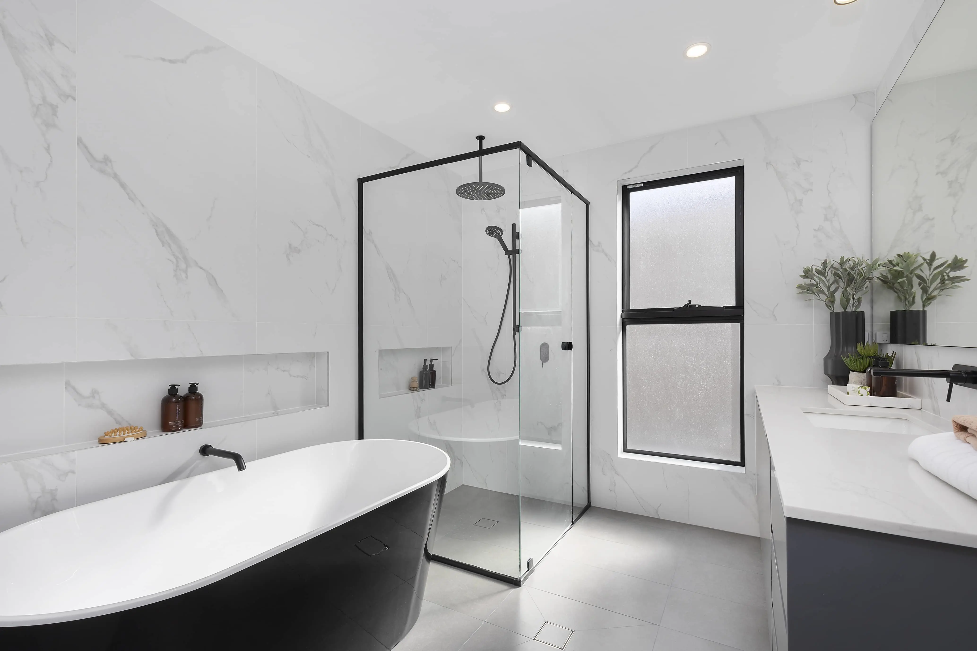 Modern bathroom with white marble walls, black-framed glass shower, freestanding bathtub, and minimalist vanity with plants.