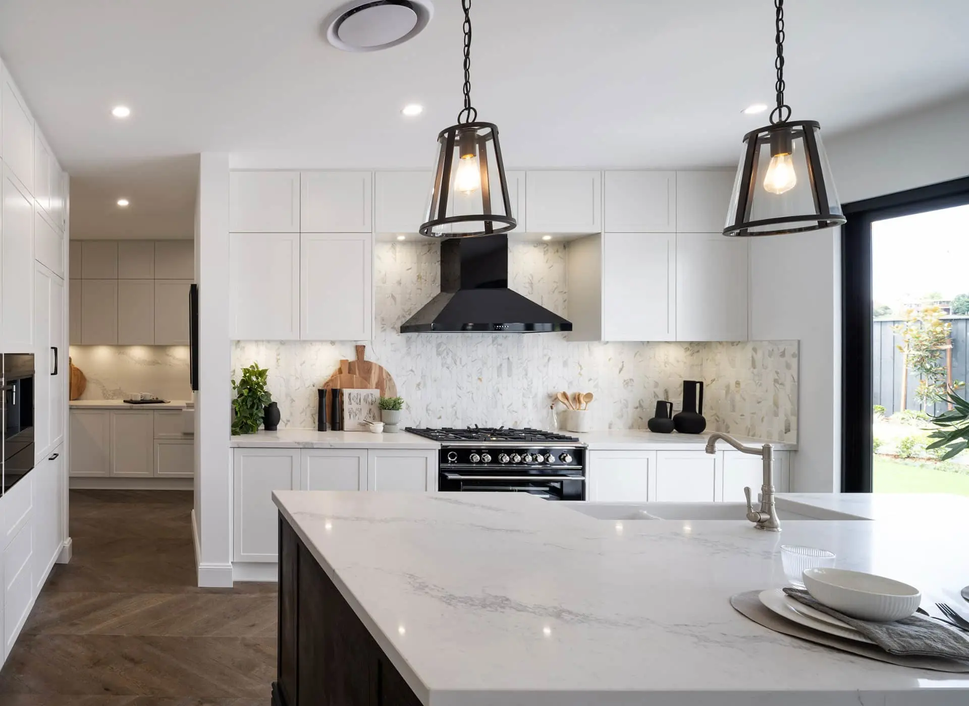 Modern white kitchen with marble countertops, black range hood, pendant lights, and island with sink overlooking garden.