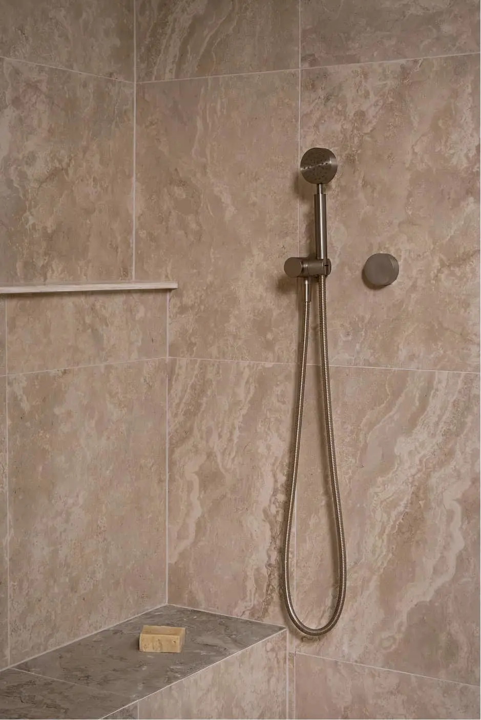 Handheld shower head with metal hose mounted on beige tiled shower wall with corner shelf and soap bar.