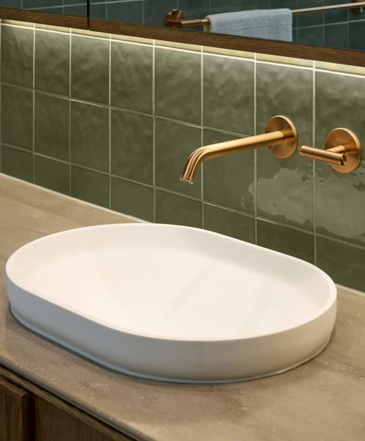 Modern bathroom with white oval sink and brass wall-mounted faucet against green tiled wall.