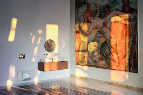 Modern bathroom with copper vanity, white sink, round mirror, and large Renaissance-style artwork on wall with warm light patterns.