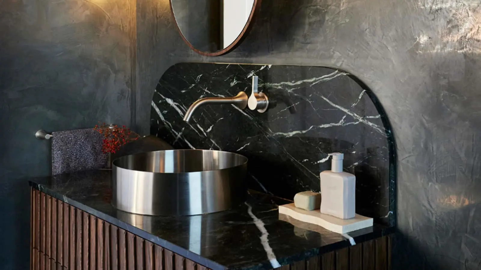 Modern bathroom with black marble sink, brass faucet, metal basin, wooden cabinet, and round mirror on dark walls.