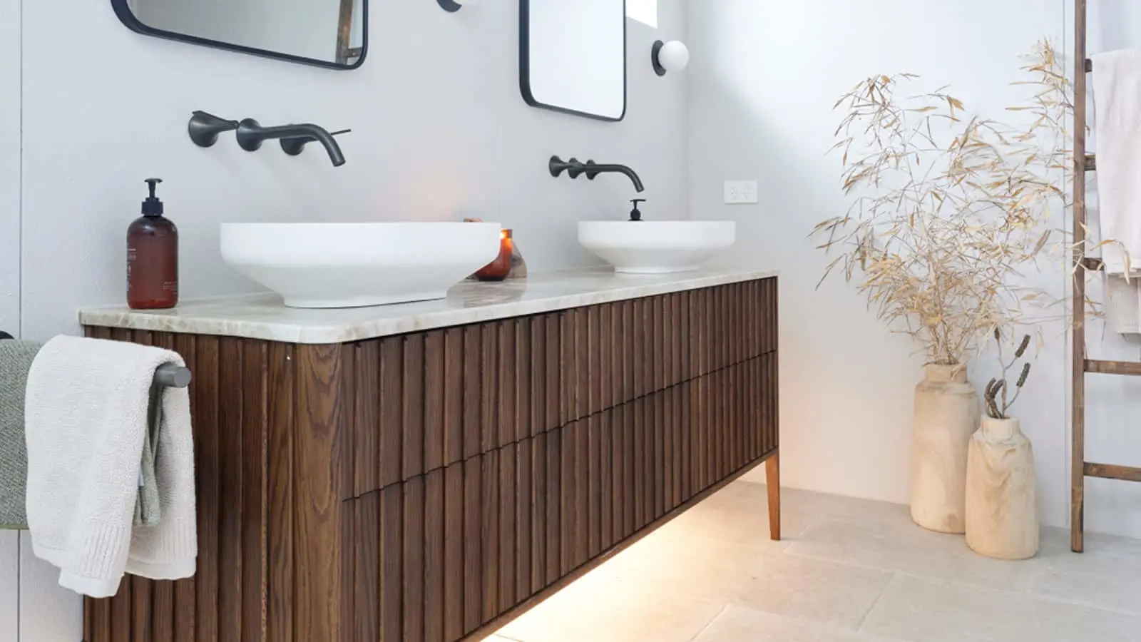 Modern bathroom with wooden vanity, two white vessel sinks, black faucets, and decorative dried plants in ceramic vases.
