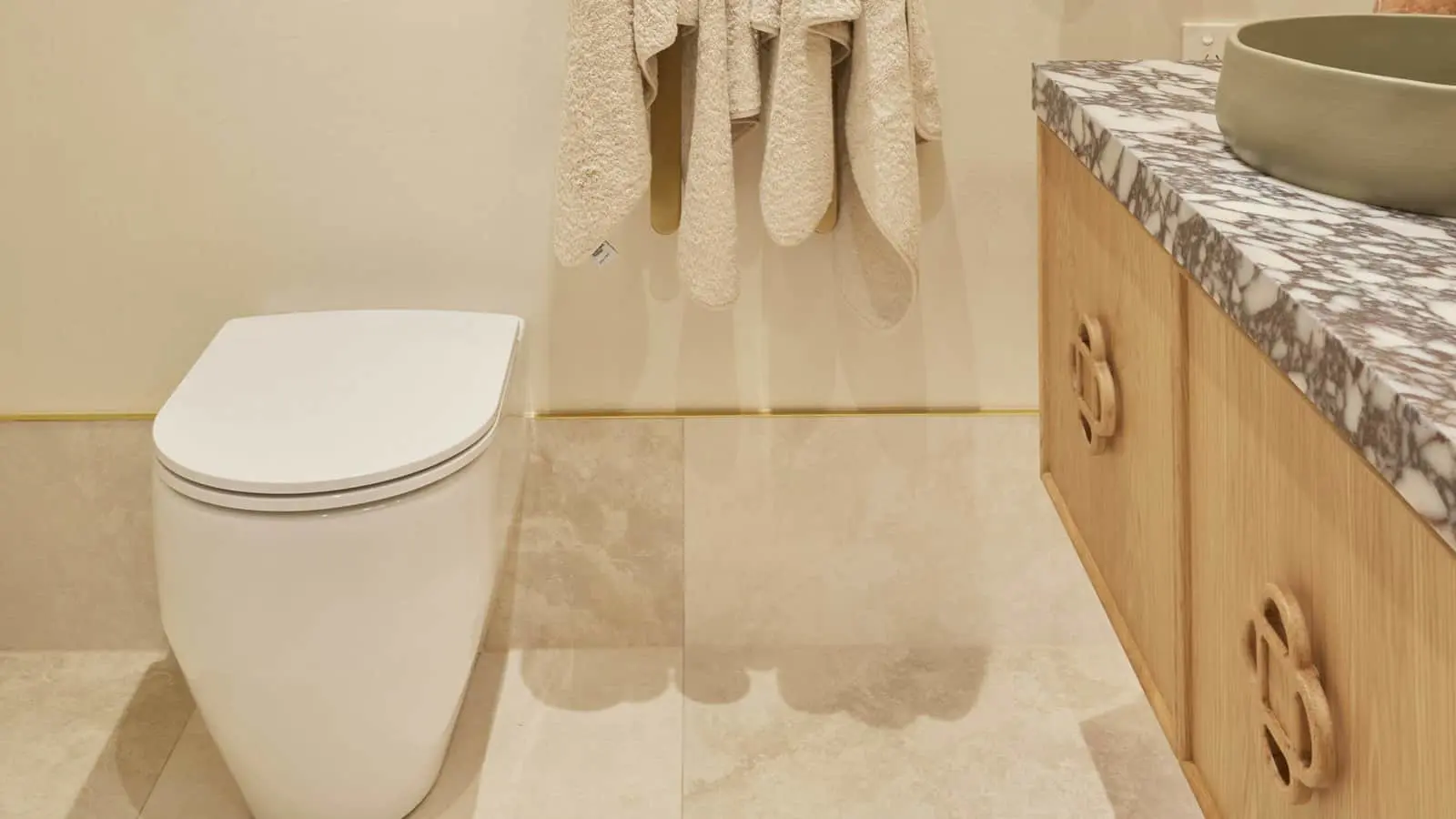 Modern bathroom with white toilet, wooden vanity with marble countertop, and cream towels hanging on the wall.