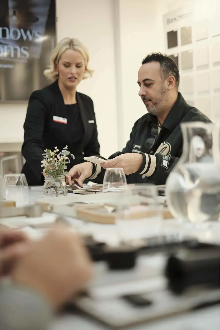 Two people in a design showroom reviewing material samples on a table with small flower arrangement and glassware.