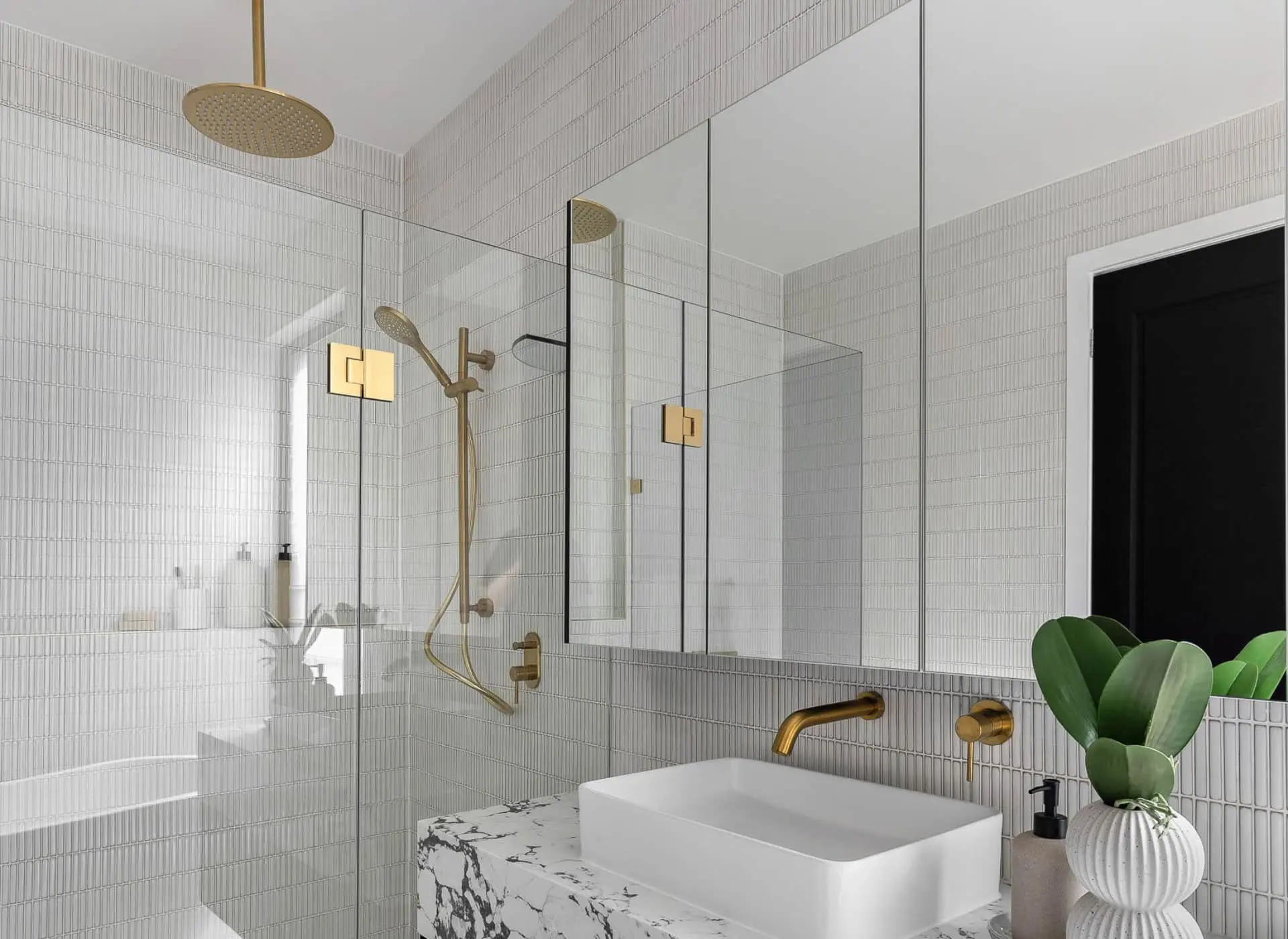 Modern white bathroom with glass shower, gold fixtures, marble countertop, rectangular sink, and potted plant.