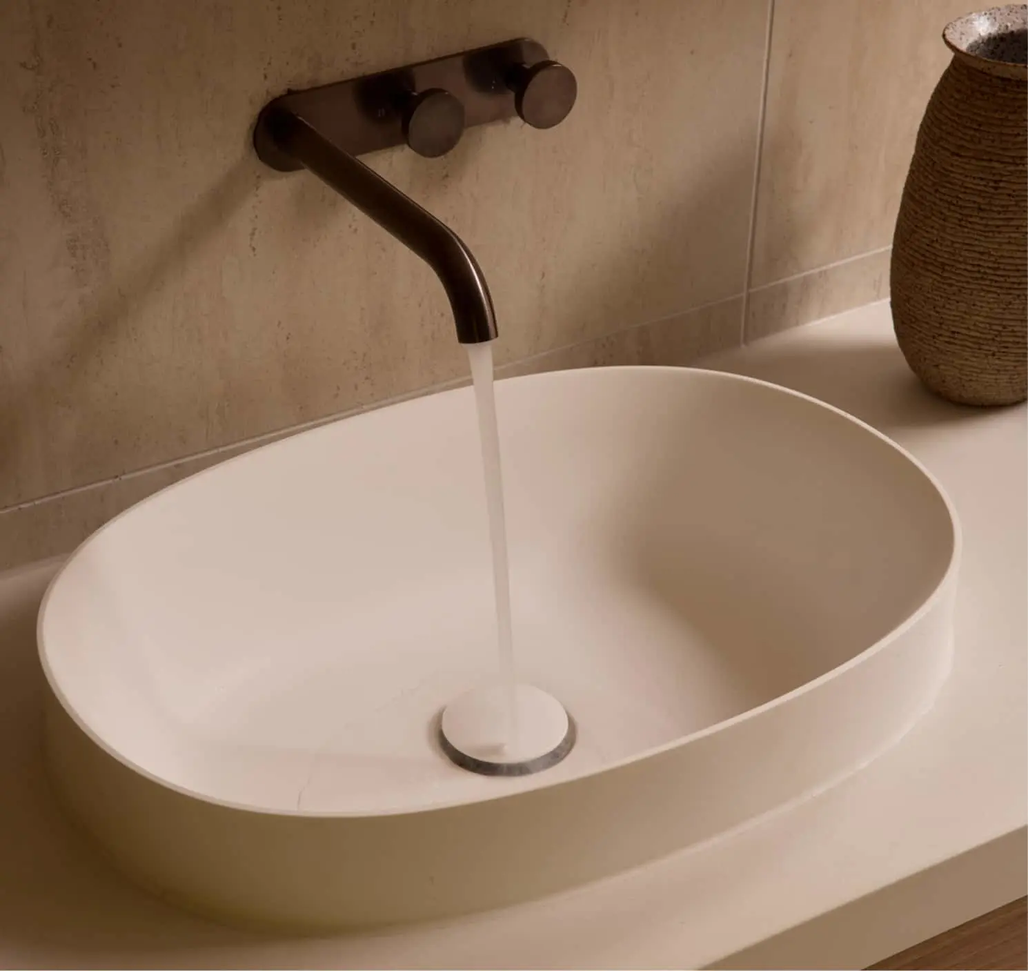 Oval white countertop basin with water running from a dark bronze wall-mounted faucet against beige tiles.