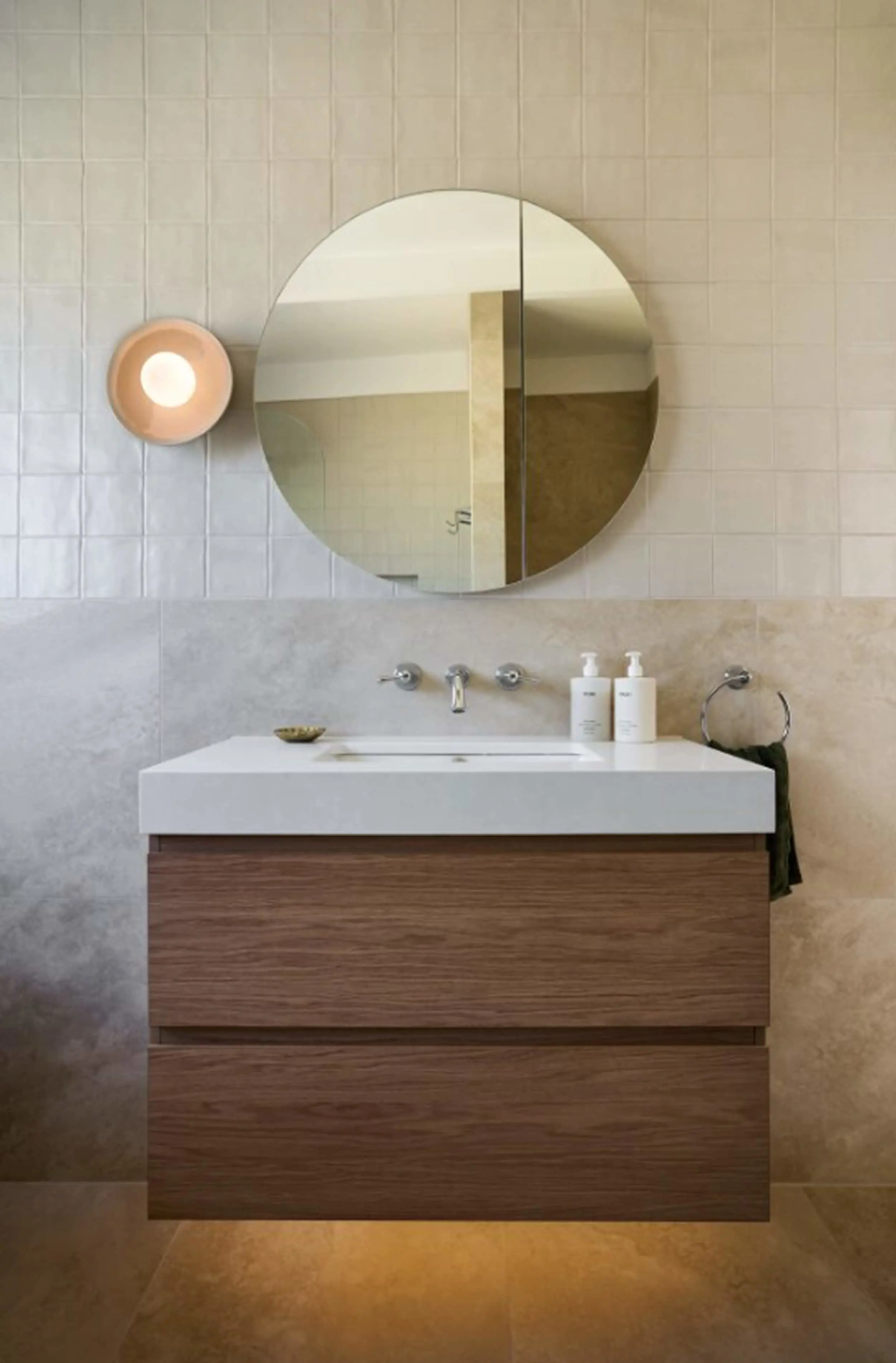 Modern bathroom with wooden vanity, white sink, round mirror, and wall sconce on tiled wall.