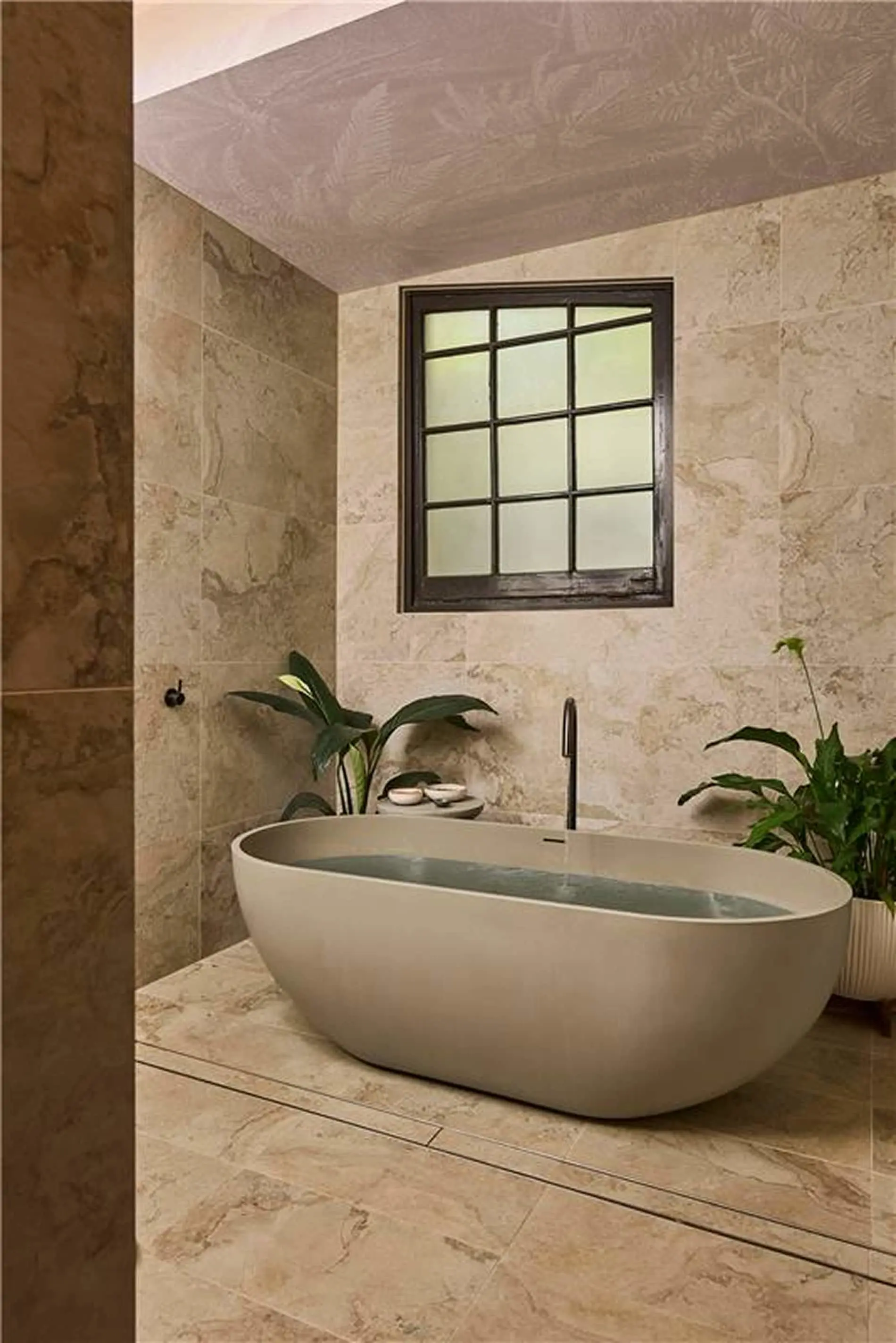 Modern bathroom with freestanding oval tub, marble tile walls, small window, and decorative plants.