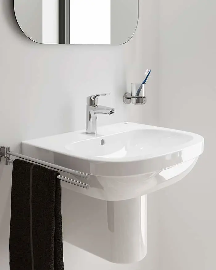 Modern white bathroom sink with chrome faucet, toothbrush holder, rounded mirror, and black towel on rail.