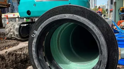 Large green pipe with black rubber seal at construction site with turquoise excavator in background.