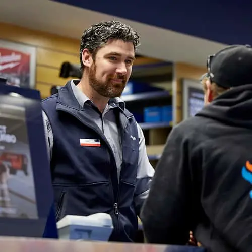 Store employee in navy blue uniform jacket speaking with a customer at a service counter in a retail environment.
