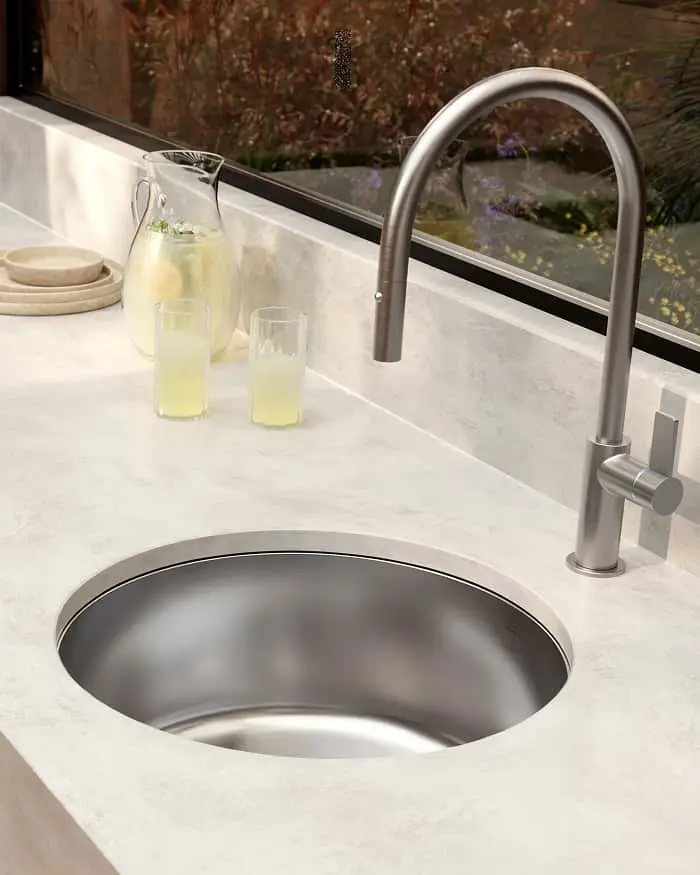 Modern kitchen sink with stainless steel faucet on white countertop, lemonade pitcher and glasses by window.