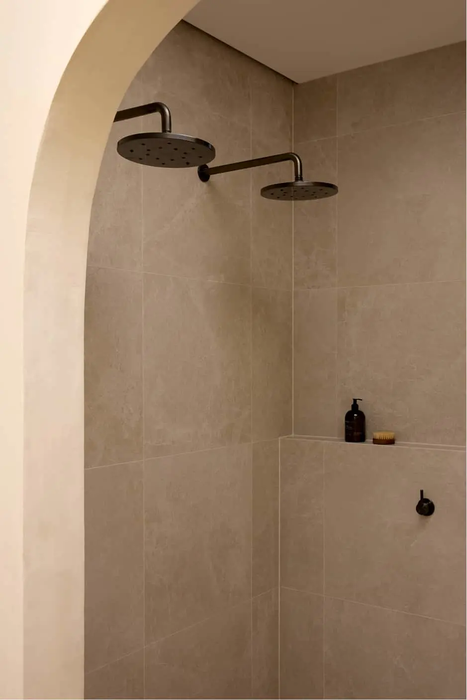 Beige tiled shower with two black rainfall showerheads and a built-in shelf holding soap dispenser and brush