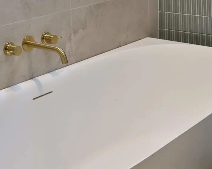 Modern white bathtub with gold wall-mounted faucet against gray tiled bathroom walls.