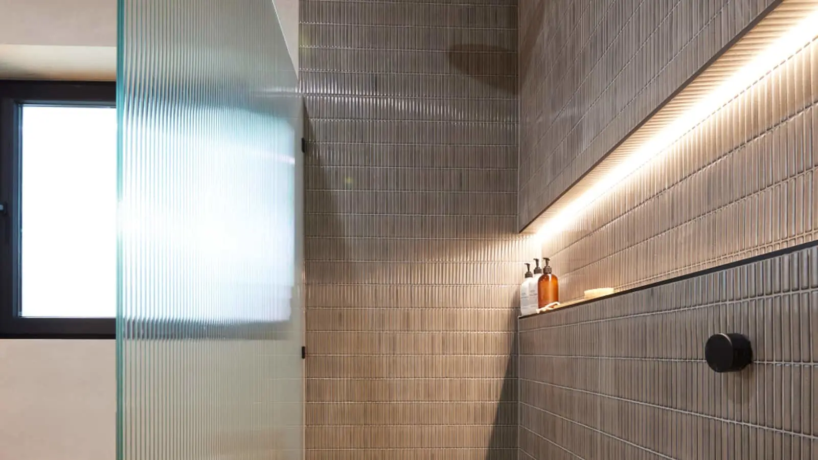 Modern bathroom with beige tiled walls, frosted glass partition, recessed lighting, and amber bottles on a shelf.