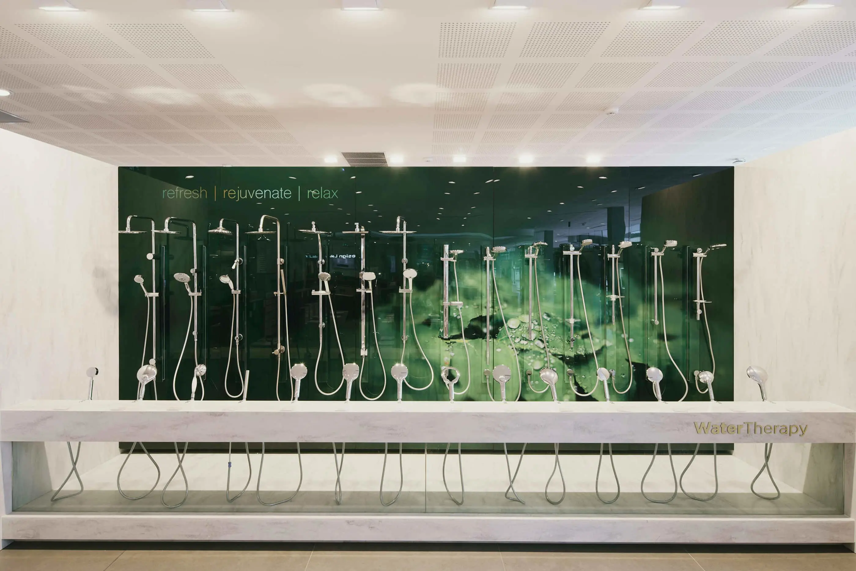 Line-up of multiple showerheads in a bathroom showroom, against a green glass wall displaying "refresh | rejuvenate | relax" text.