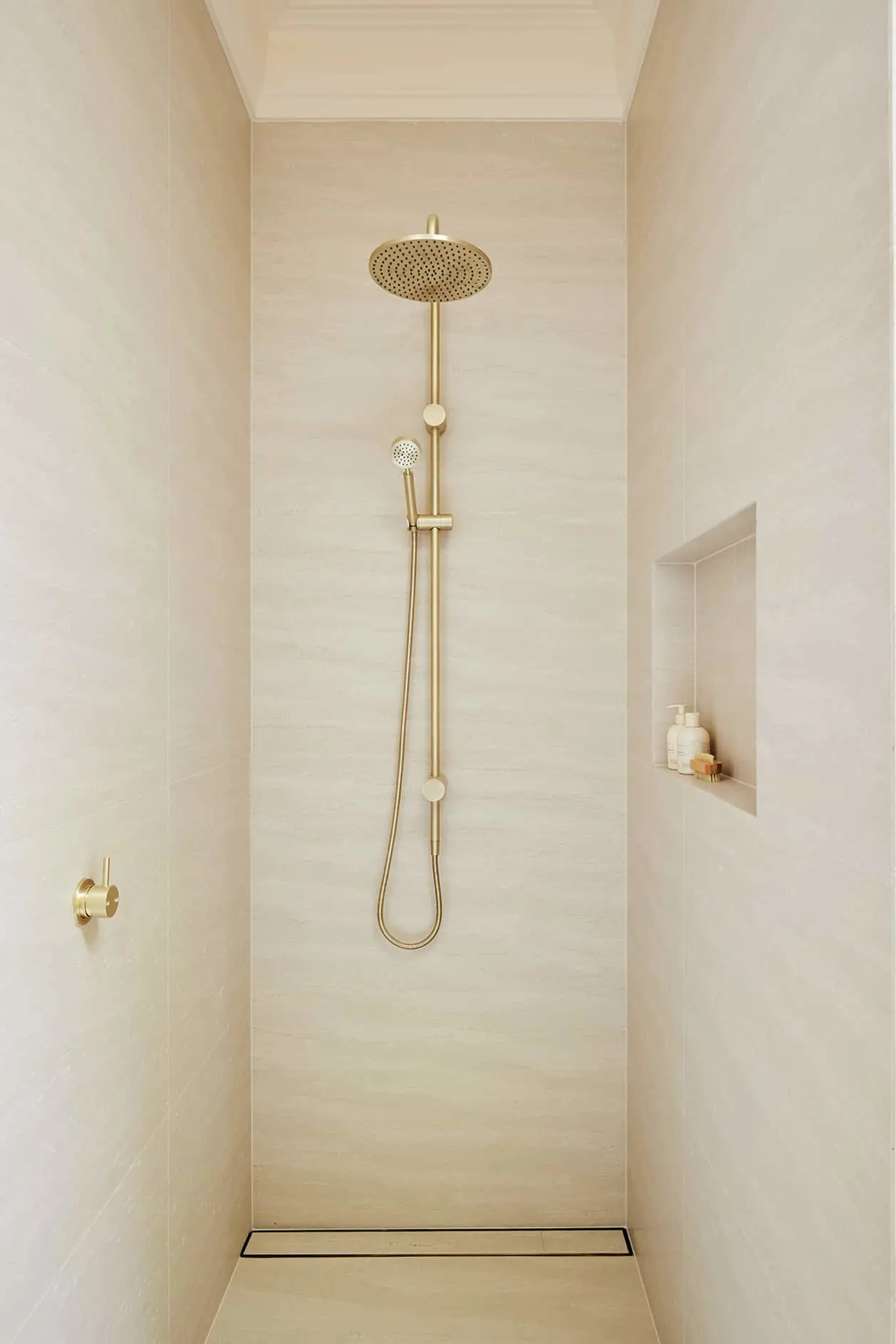 Modern shower with brass fixtures featuring rainfall showerhead and handheld nozzle in a cream-colored tiled enclosure with built-in shelf.