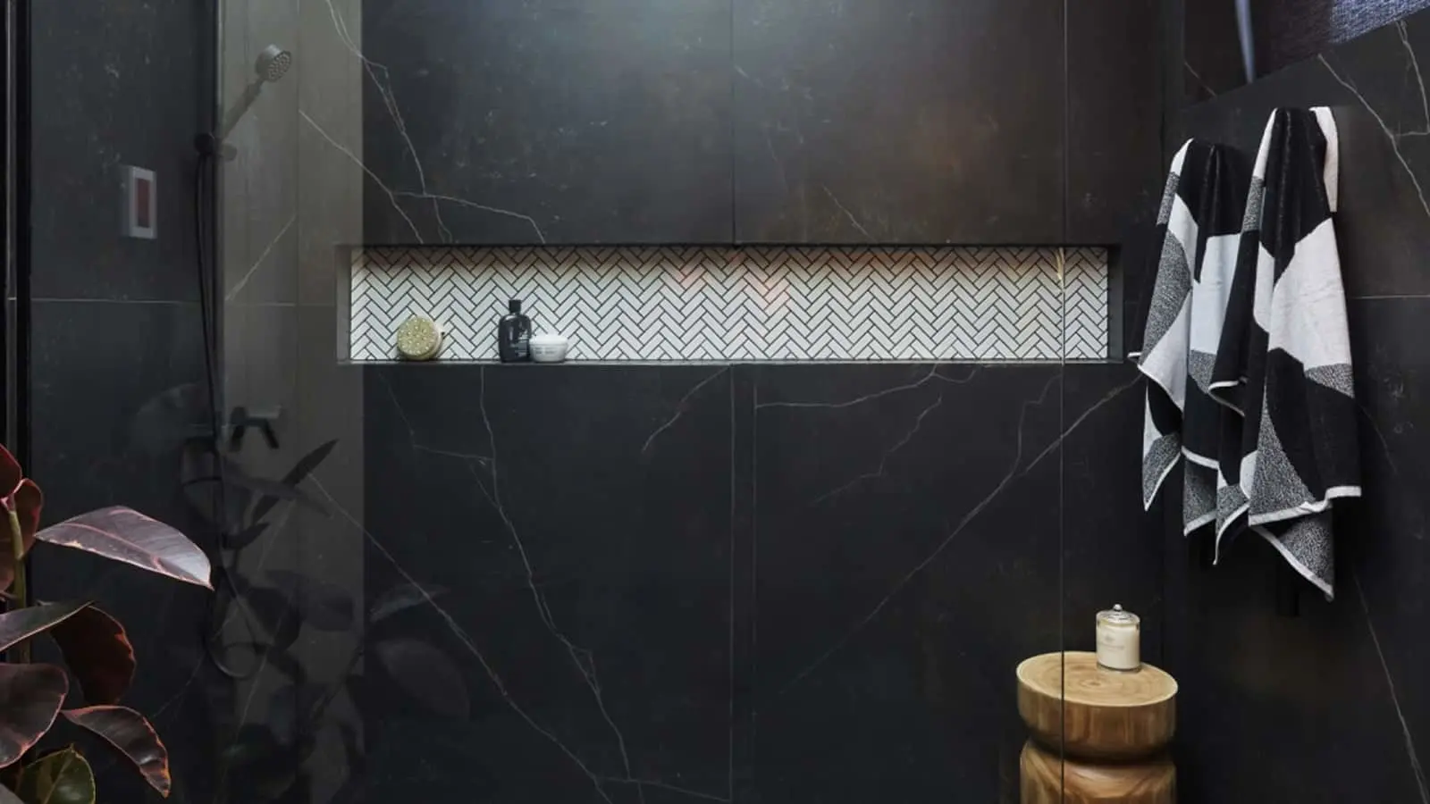 Modern bathroom with black marble walls, herringbone tile niche, black and white towel, and wooden stool with candle.