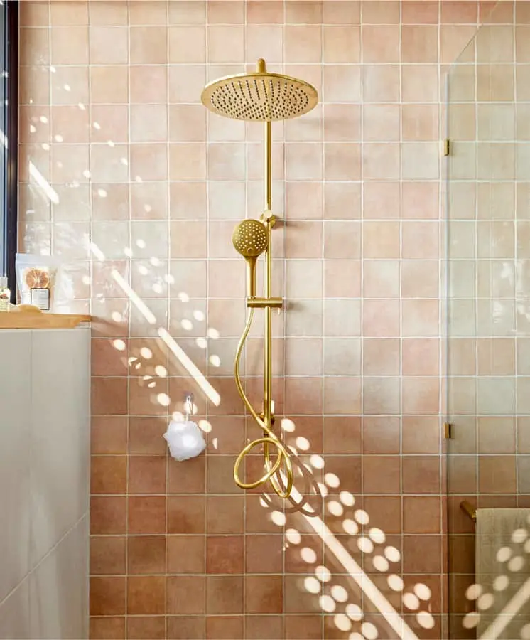 Gold rainfall shower head mounted on pink tiled bathroom wall with sunlight creating spotted light patterns.