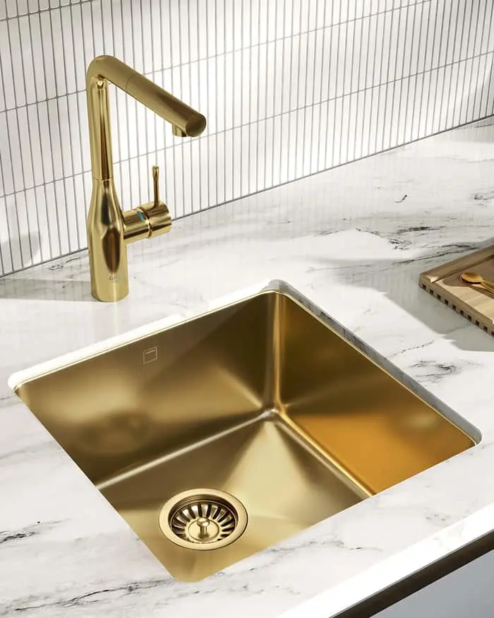 Gold kitchen sink and matching faucet installed in white marble countertop with vertical tile backsplash.