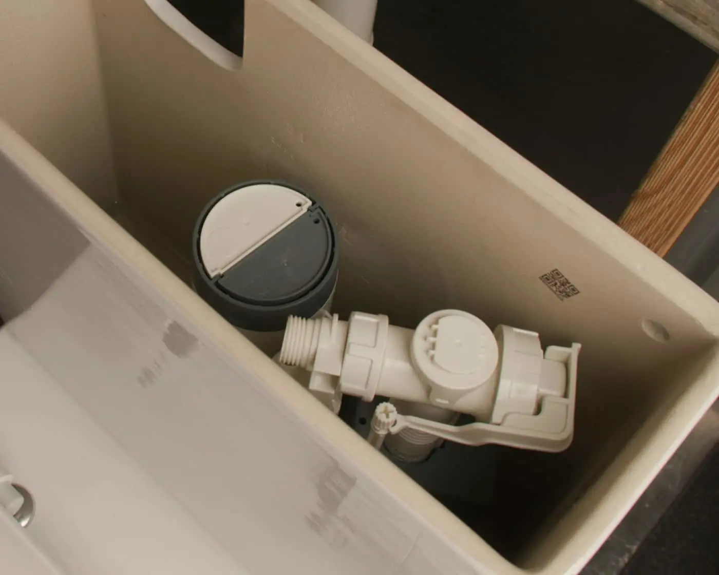 Inside of a toilet tank showing the flush valve mechanism with black and white components and plumbing fittings.