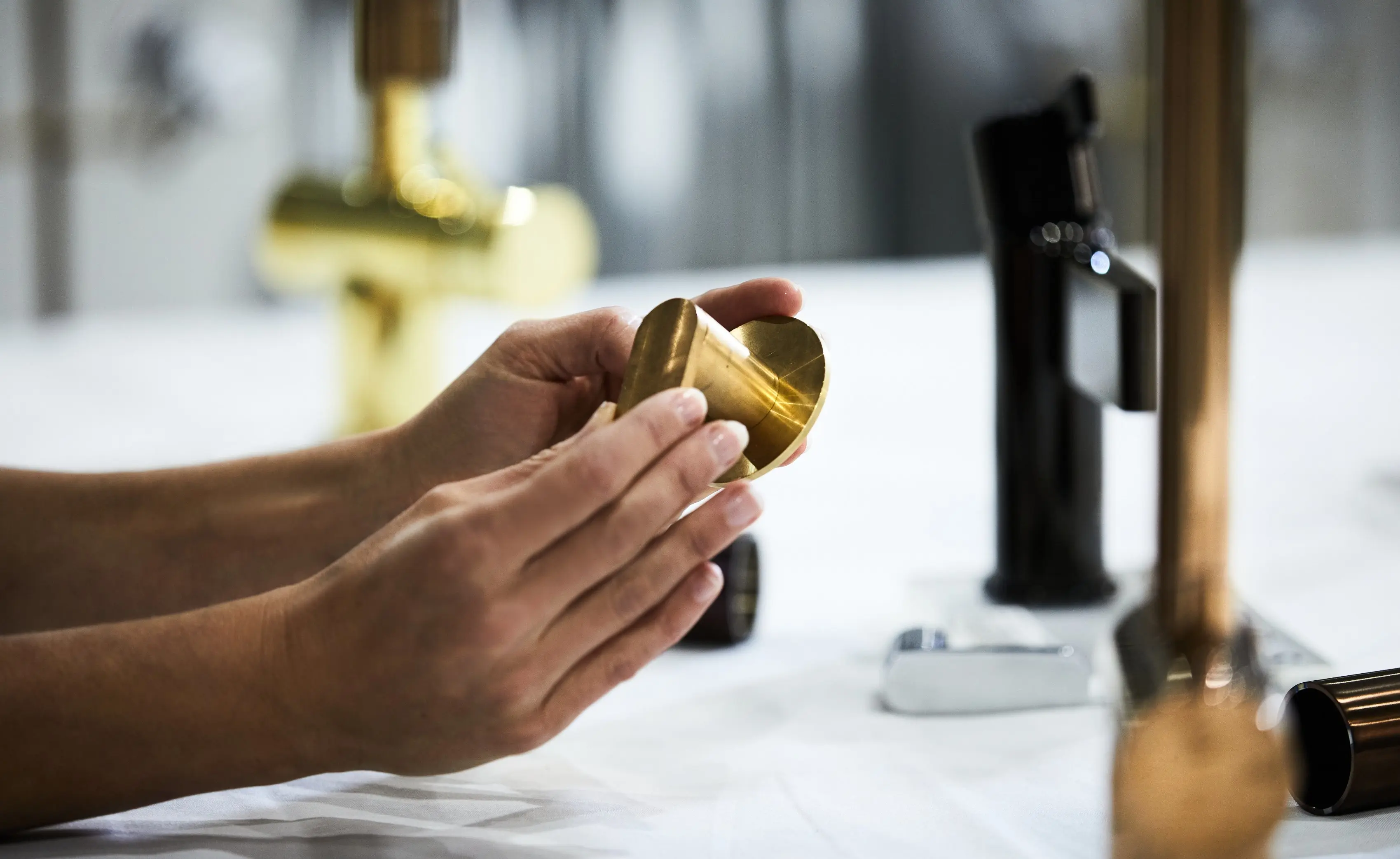 Hands holding a small brass cylindrical component, with various faucet fixtures on a white surface in the background.