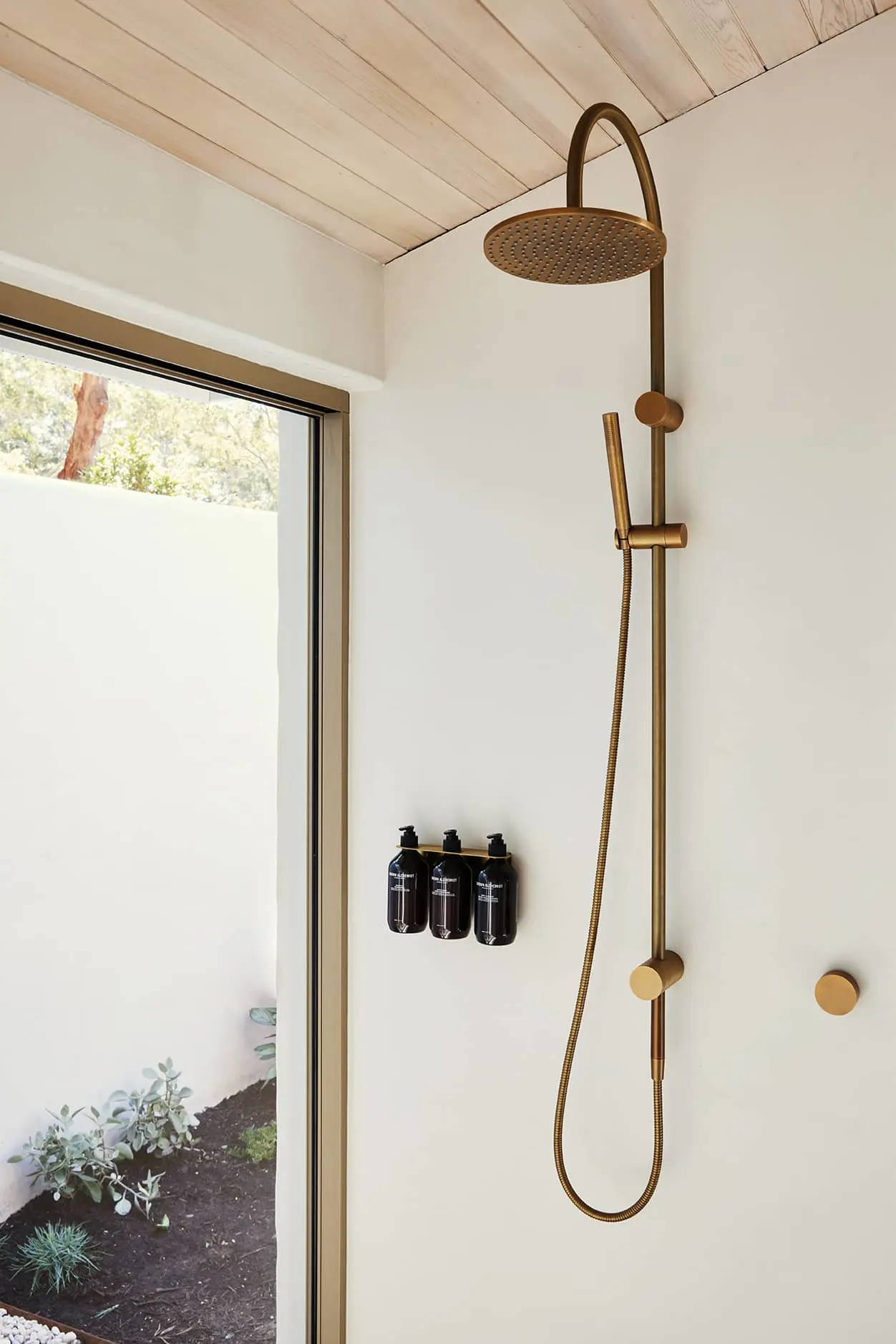 Modern brass shower fixture with rainhead and handheld sprayer in white bathroom with window view to garden.