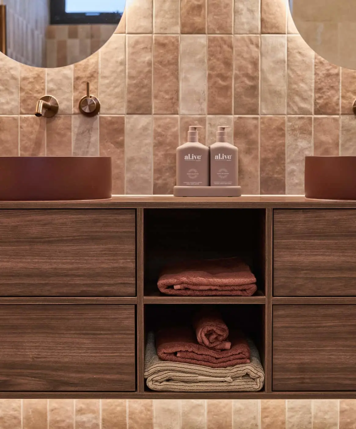 Modern bathroom vanity with dark wood cabinets, beige tile walls, maroon basins, and neatly folded towels.