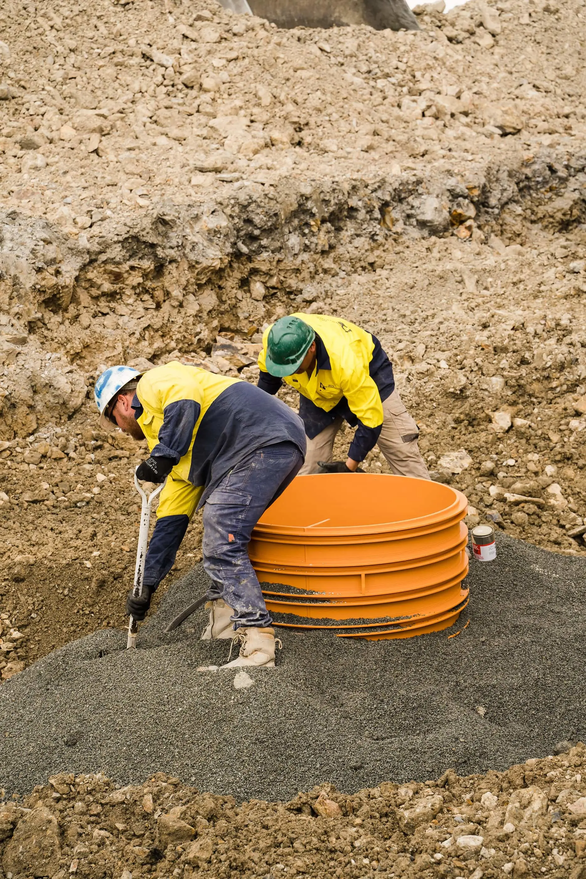 Construction workers in yellow safety vests installing orange drainage pipes in excavated soil.