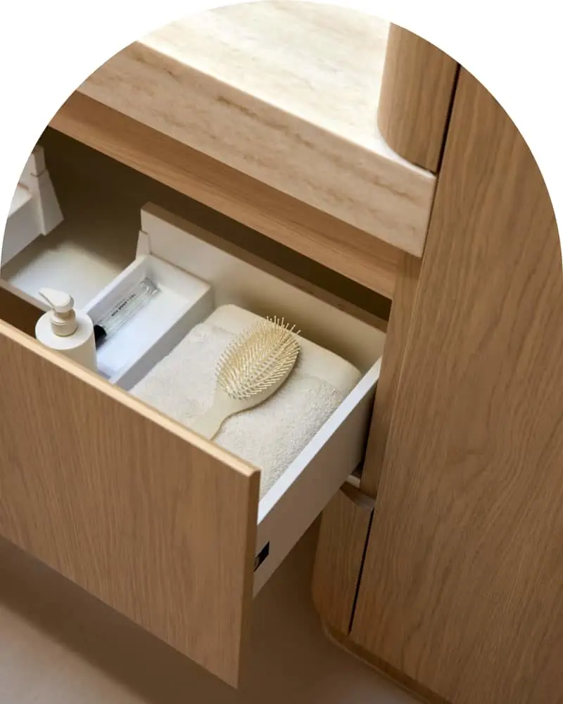 Light wood bathroom cabinet with an open drawer containing a folded towel, hairbrush, pump bottle, and small white boxes.