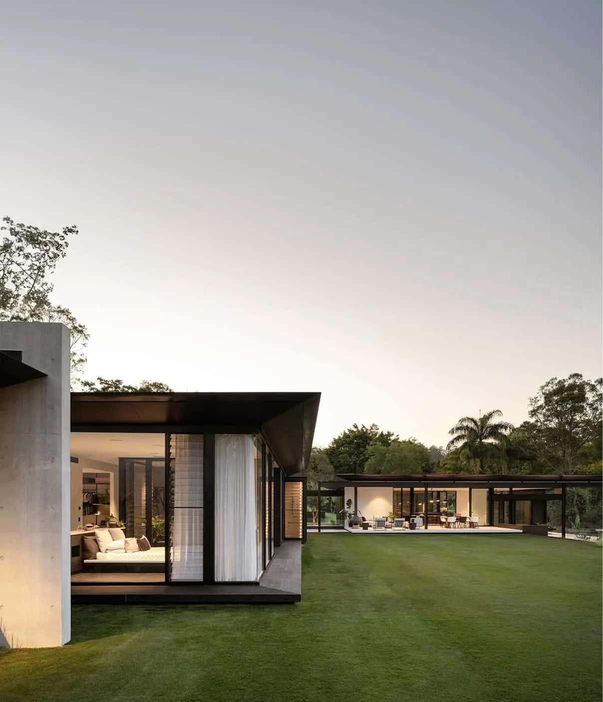 Modern L-shaped house with glass walls, concrete elements, and manicured lawn, set against twilight sky and tropical trees.