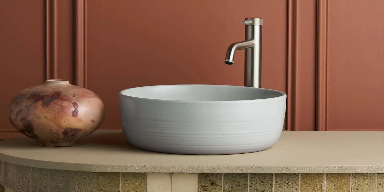 Modern white vessel sink with brushed nickel faucet next to decorative ceramic vase against terracotta wall.