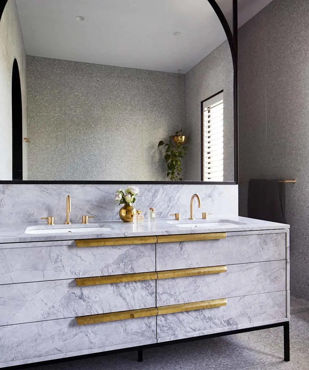 Modern bathroom with marble vanity, brass fixtures, double sinks, large mirror, and gray textured walls.