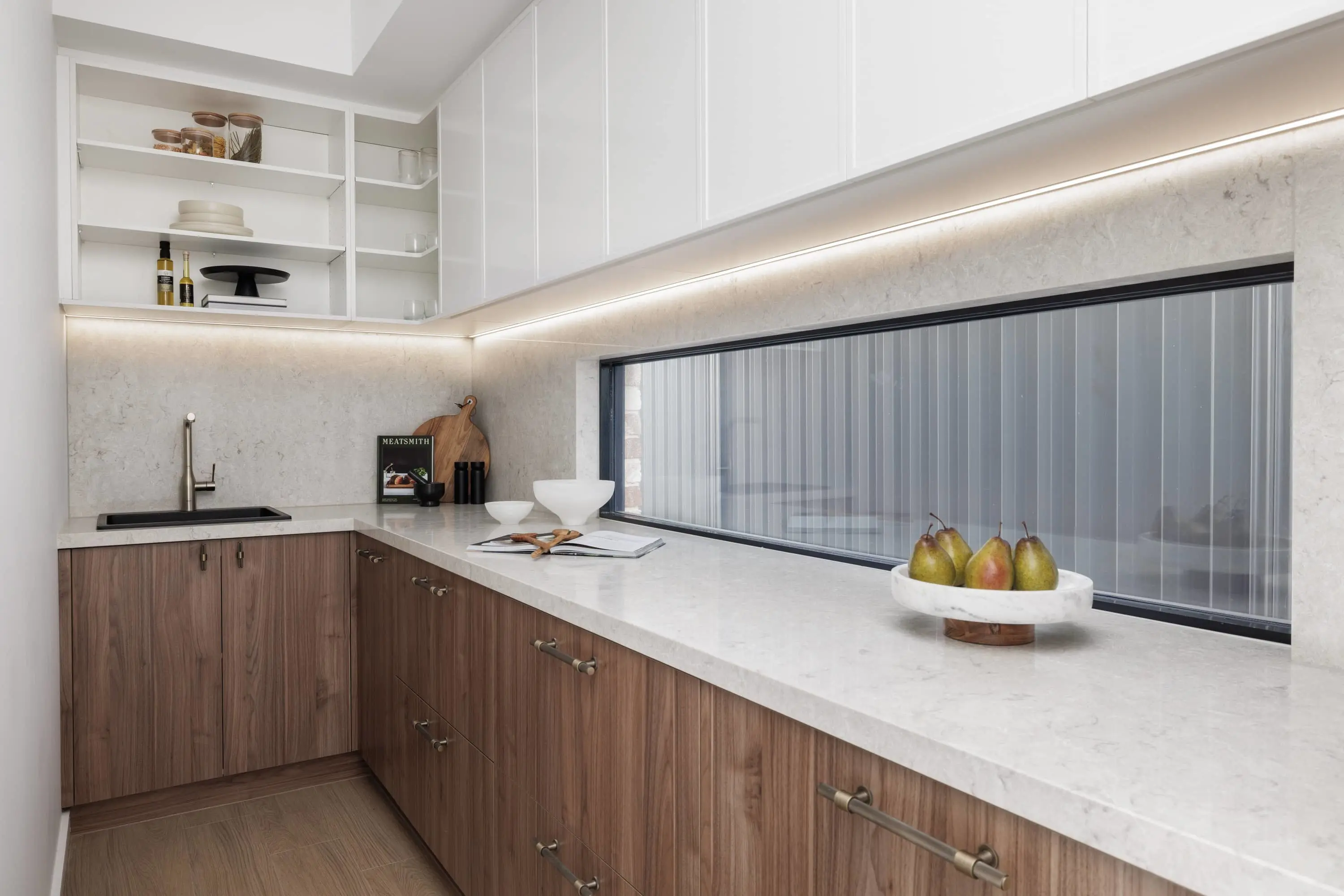 Modern kitchen with white upper cabinets, wooden lower cabinets, marble countertops, and pears in a white dish by a window.