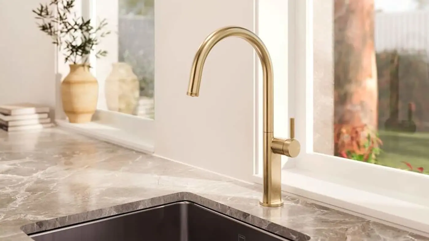 Modern gold kitchen faucet on stone countertop with dark sink, decorative vases and window view in background.
