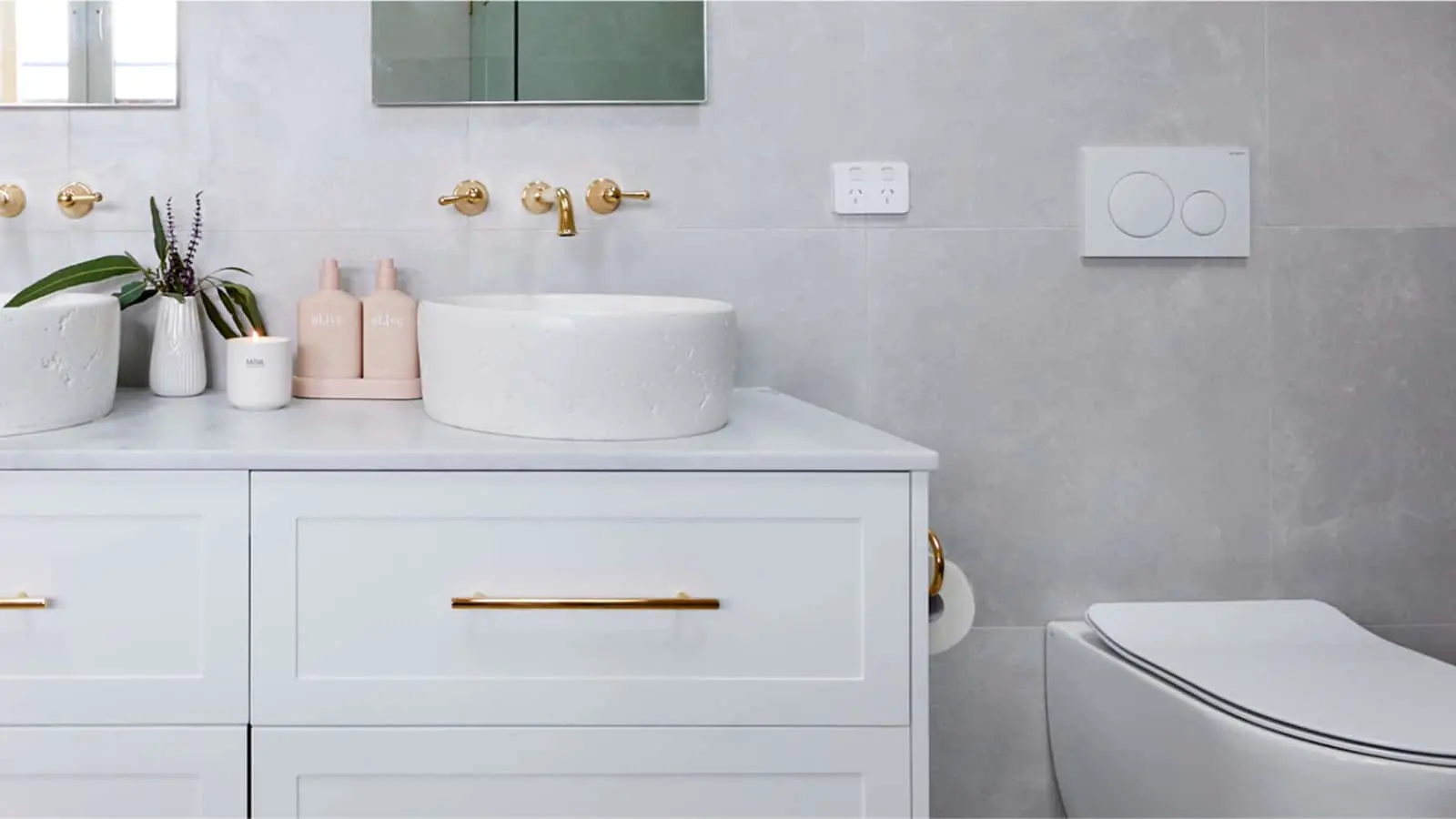 Modern white bathroom with round vessel sink, gold fixtures, pink toiletry bottles, and plant on white vanity.