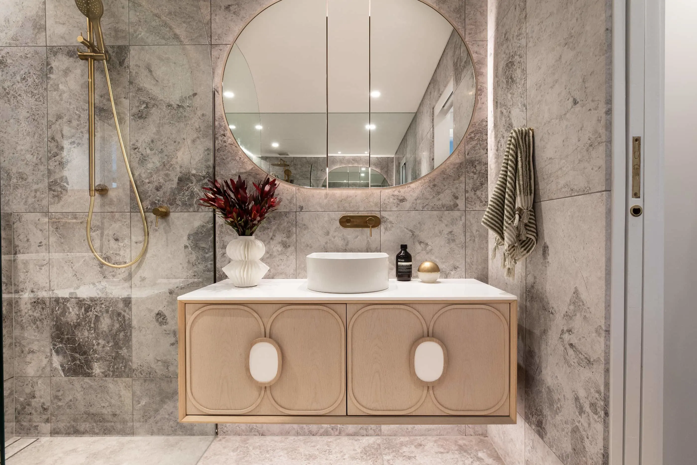 Modern bathroom with marble walls, round mirror, wooden vanity, white vessel sink, gold fixtures, and red flowers in vase.