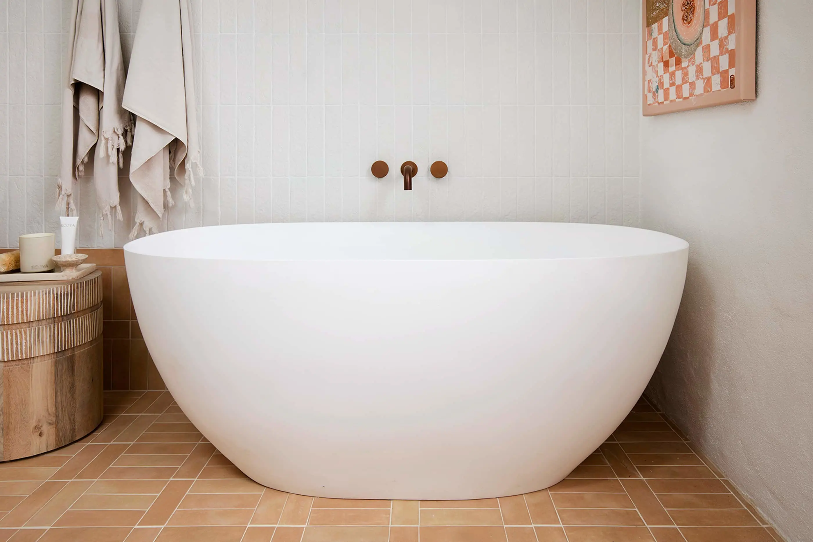 Modern white freestanding bathtub with brass wall-mounted faucet, beige towel hanging nearby, and wooden side table.
