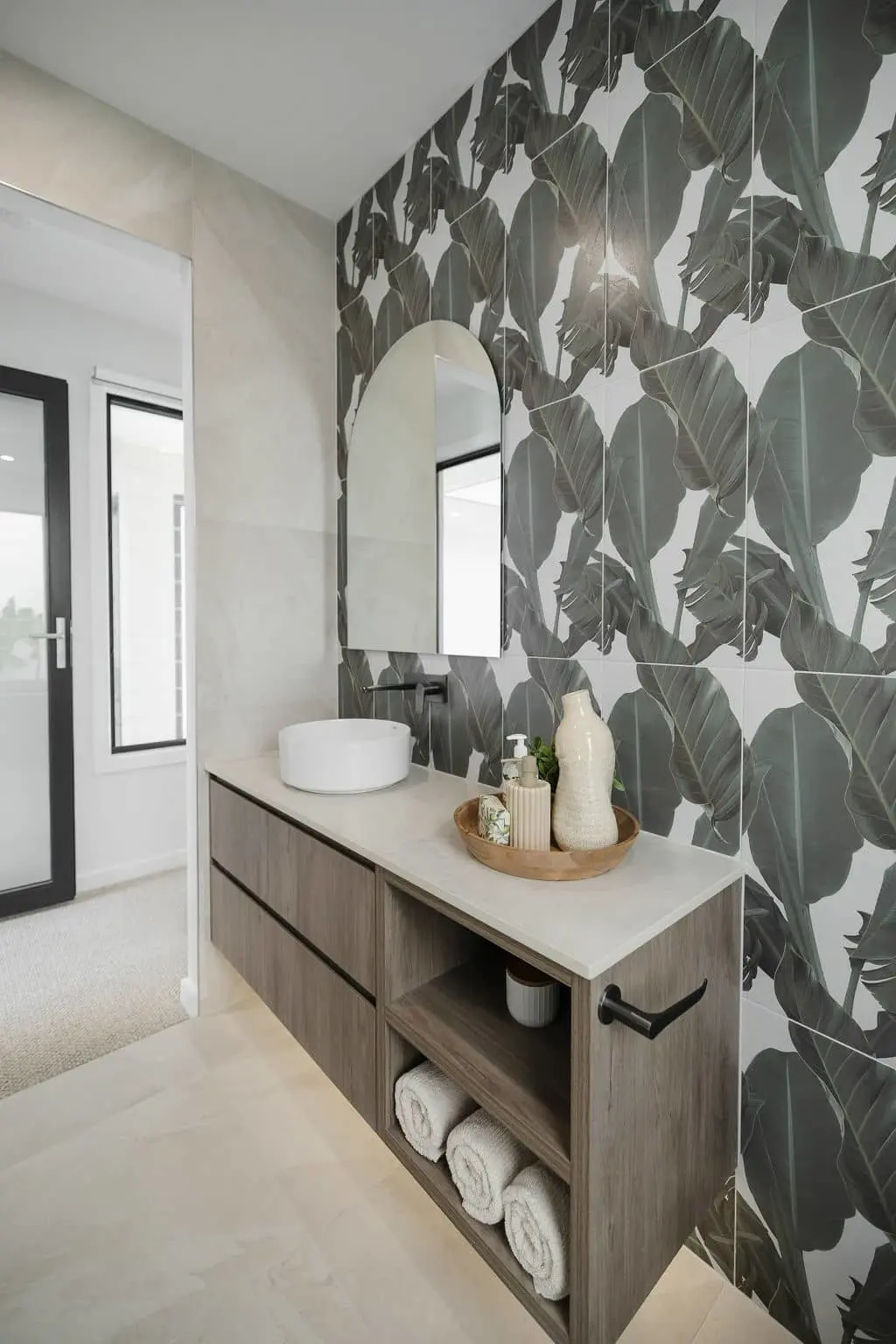 Modern bathroom with tropical leaf wallpaper, wooden vanity, white vessel sink, and oval mirror.