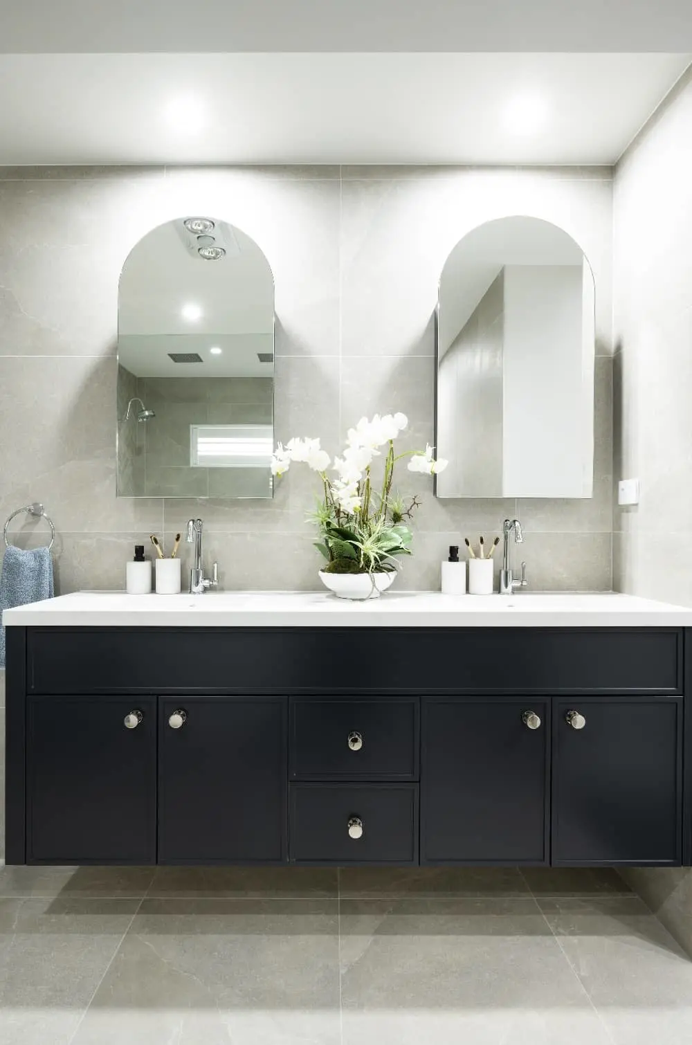 Modern bathroom with navy vanity, white countertop, dual sinks, arched mirrors, and white orchid centerpiece.