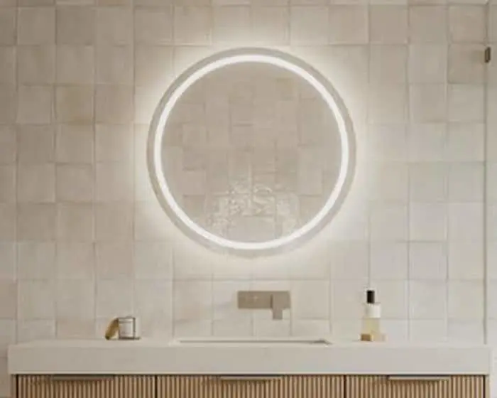 Circular illuminated mirror above white bathroom vanity with wooden slatted cabinet and white tile backsplash.