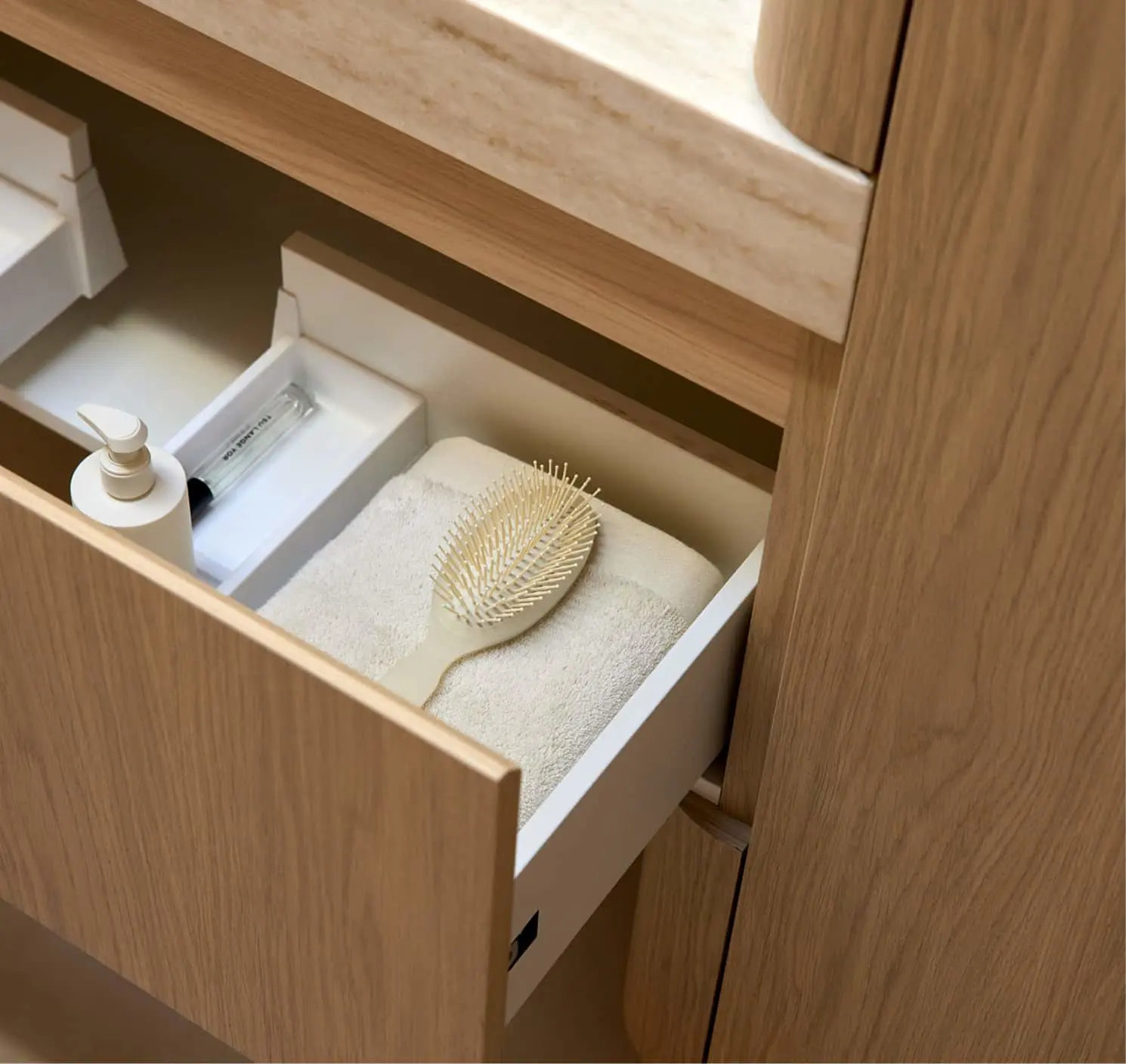Open light wood bathroom drawer containing a folded cream towel, hairbrush, pump bottle, and small white boxes.
