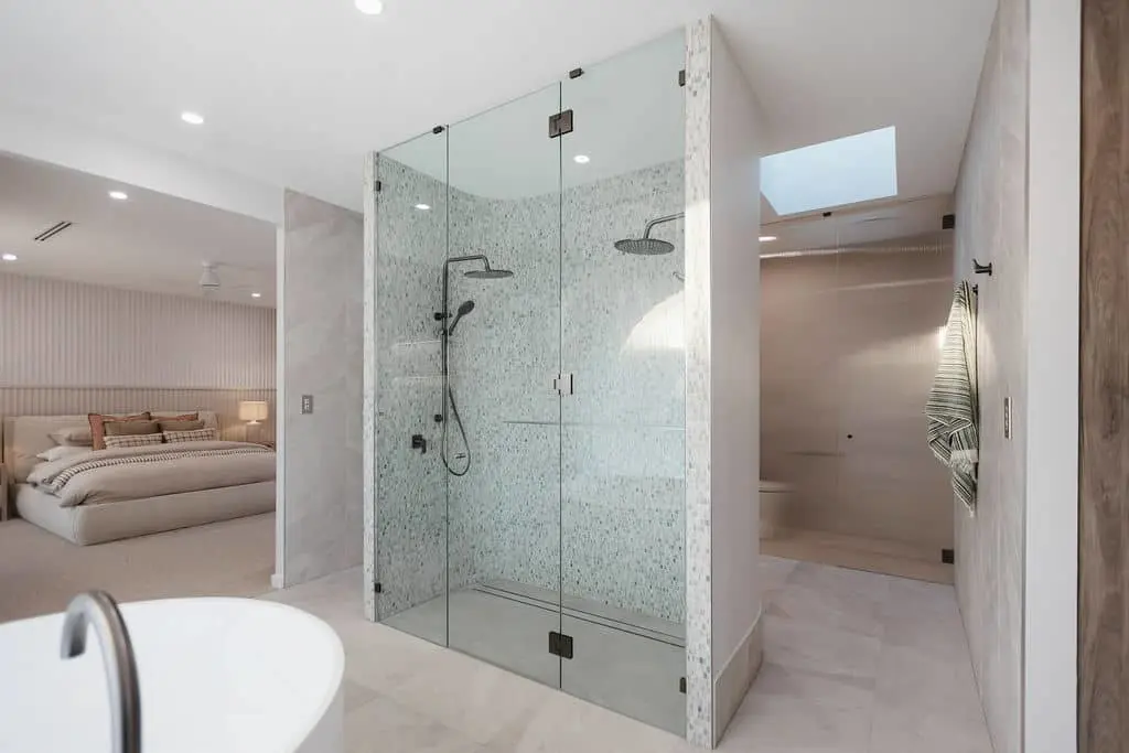 Modern bathroom with glass shower enclosure featuring mosaic tile walls, connected to a bedroom with neutral decor.