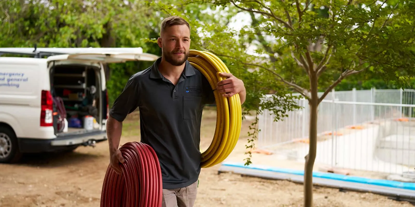 Tradesperson in dark polo carrying yellow and red pipes at a worksite with service van in background.