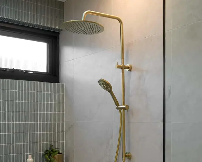 Modern bathroom with gold rainfall shower system mounted on light marble tile wall, small window above tiled half-wall.