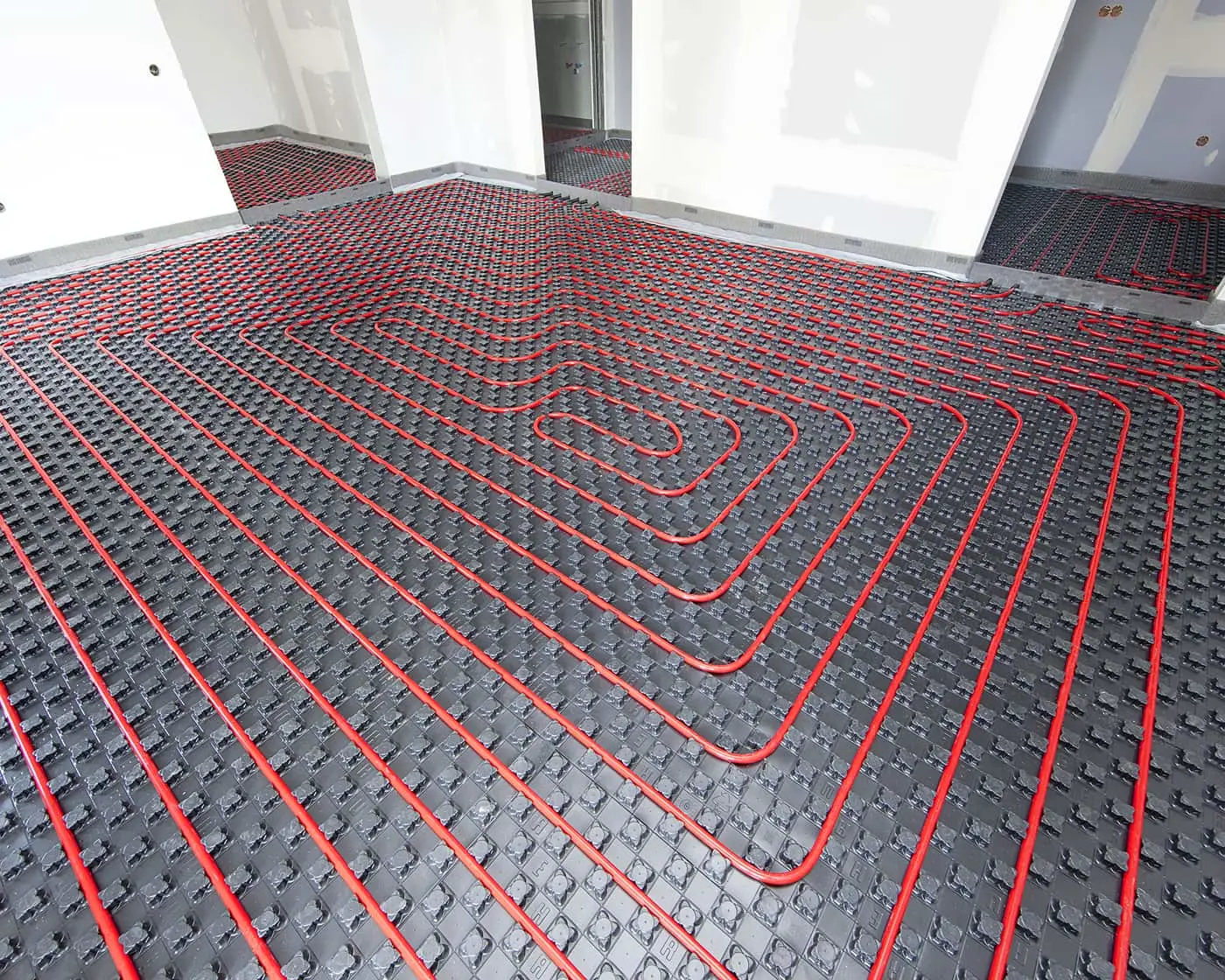 Underfloor heating installation with red pipes arranged in loops across black insulation panels in a room under construction.